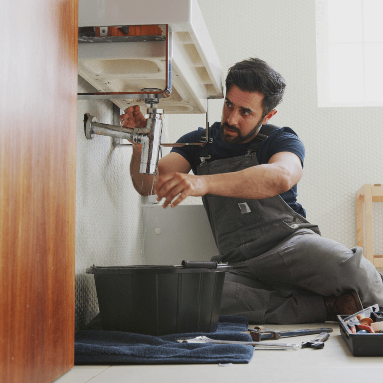 Man in overalls kneels under a sink, tightening exposed plumbing with tools nearby in a tiled room
