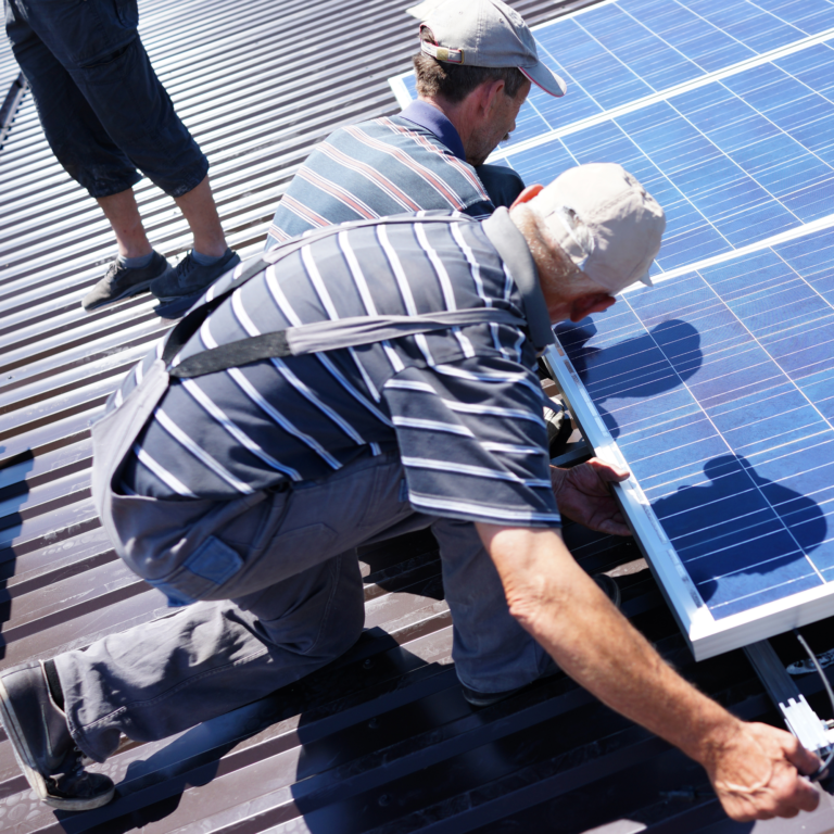 Two workers install blue solar panels on a corrugated metal roof, kneeling and handling a panel together