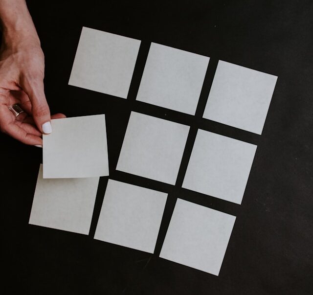 Hand picks up a white square card from a 3x3 grid of blank squares on a black table.